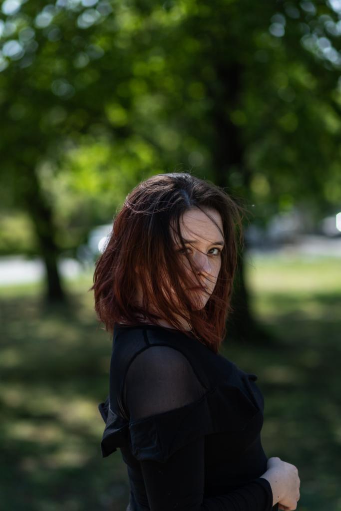 Portret kobiety z rudymi włosami w czarnej bluzce z prześwitującymi rękawami, patrzącej przez kosmyki włosów na tle zieleni drzew w plenerze.