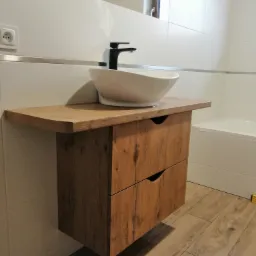 Wisząca szafka łazienkowa z drewnopodobnymi frontami, umywalka nablatowa w kształcie misy, czarna bateria, białe płytki na ścianie i podłoga imitująca drewno.