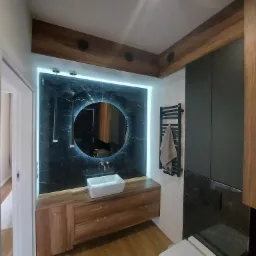 Nowoczesna łazienka z podświetlanym lustrem, drewnianymi szafkami pod umywalką i czarnym marmurem na ścianach, widoczny grzejnik drabinkowy z ręcznikiem i toaleta.