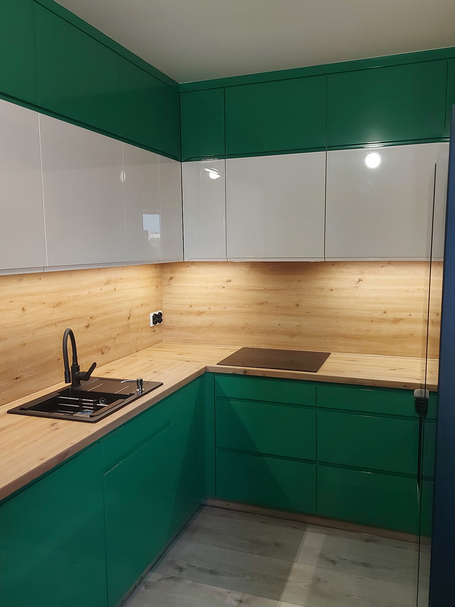 Nowoczesna kuchnia z zielonymi szafkami dolnymi i białymi górnymi, drewniany blat, czarny zlew i bateria, oświetlenie podszafkowe