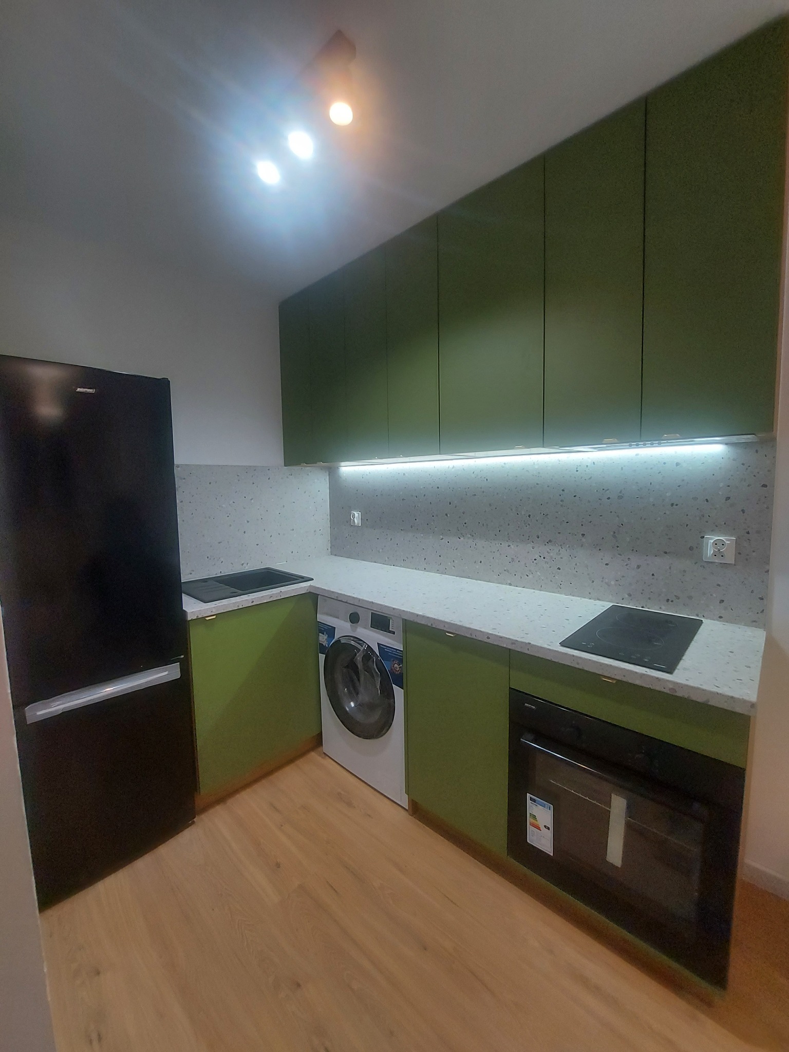 Nowoczesna kuchnia w odcieniach zieleni z czarnym sprzętem AGD: lodówka i piekarnik. Blat z motywem lastryko, oświetlenie LED pod szafkami wiszącymi, pralka w zabudowie.