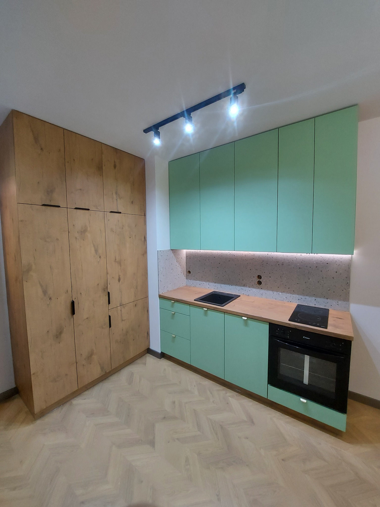 Nowoczesna kuchnia z miętową zabudową, drewnianym blatem i szafą w kolorze drewna, oświetlona listwą LED pod szafkami wiszącymi i reflektorami sufitowymi.