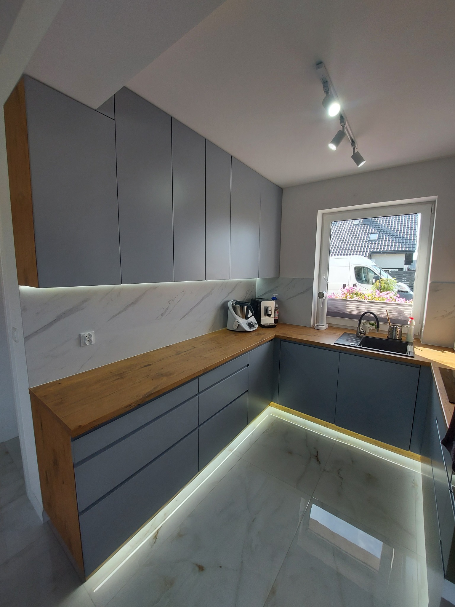 Nowoczesna kuchnia w szarościach z drewnianym blatem, oświetleniem LED pod szafkami i marmurową ścianą, widok na podjazd z samochodem dostawczym przez okno.