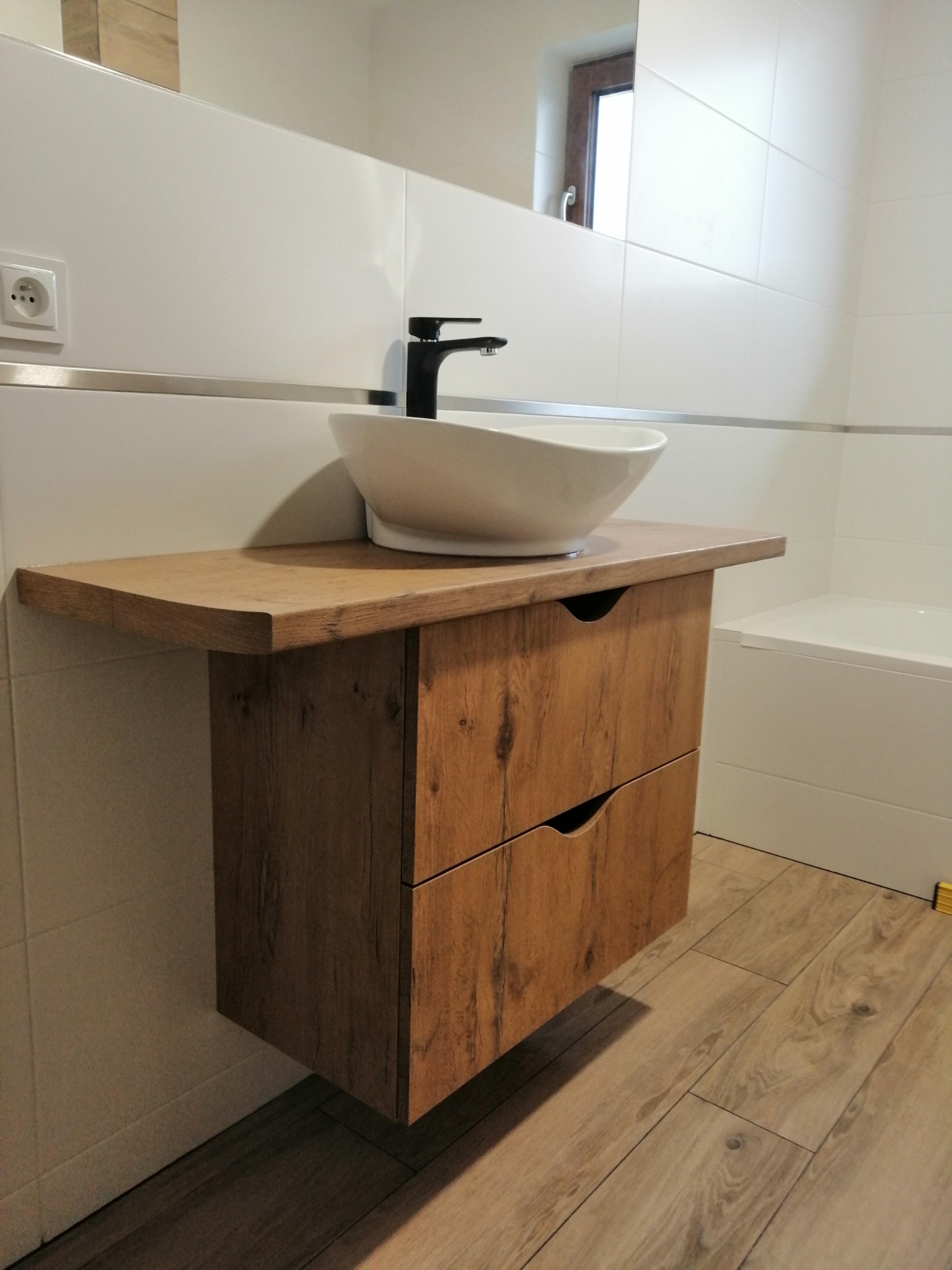 Wisząca szafka łazienkowa z drewnopodobnymi frontami, umywalka nablatowa w kształcie misy, czarna bateria, białe płytki na ścianie i podłoga imitująca drewno.