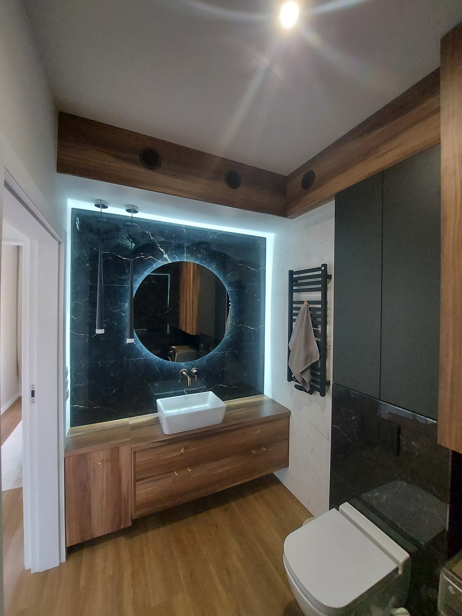 Nowoczesna łazienka z podświetlanym lustrem, drewnianymi szafkami pod umywalką i czarnym marmurem na ścianach, widoczny grzejnik drabinkowy z ręcznikiem i toaleta.