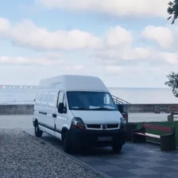 Biały bus Renault Master z numerem telefonu na boku zaparkowany na tle morza i plaży w pogodny dzień.