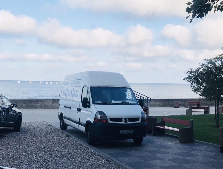 Biały bus Renault Master z numerem telefonu na boku zaparkowany na tle morza i plaży w pogodny dzień.