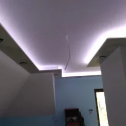 Sufit podwieszany z oświetleniem LED w kształcie litery L, widoczne kable zwisające z sufitu, ściany w odcieniach bieli i błękitu, widoczny fragment pokoju z meblami.