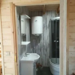 Wnętrze łazienki w drewnianym domu: kabina prysznicowa z zaokrąglonymi drzwiami, umywalka z szafką, lustro z półką i bojler na ścianie wyłożonej panelami imitującymi drewno.