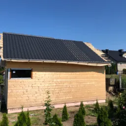 Surowy, drewniany budynek z czarnym dachem z blachy trapezowej w trakcie budowy, widoczne ocieplenie fundament&oacute;w i niezagospodarowany teren.