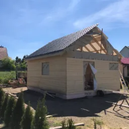 Surowa konstrukcja domu z drewnianych bali z widoczną izolacją dachu i folią ochronną, częściowo pokryta dachówką, na tle błękitnego nieba i zieleni przydomowego ogrodu.