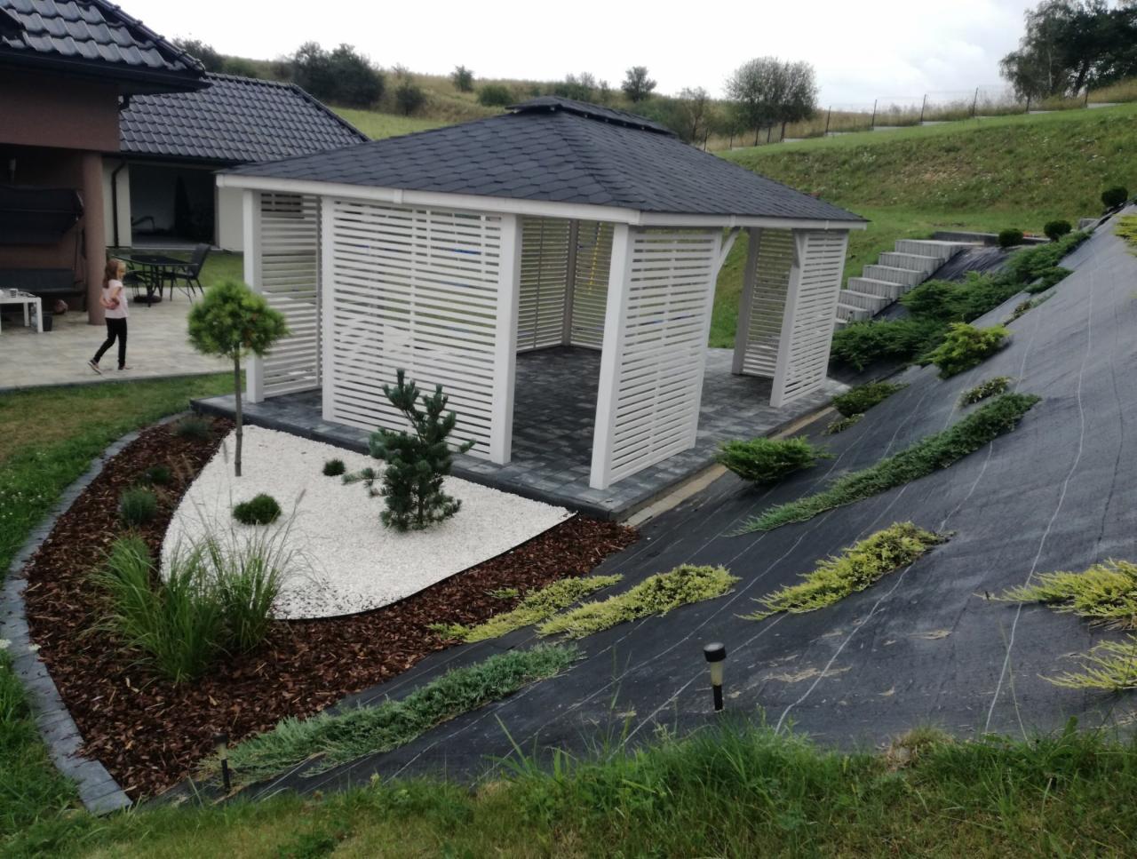 Biała, ażurowa altana z ciemnym dachem, otoczona ozdobnym ogrodem z kamieniami i korą, na tle zielonego wzgórza z roślinami okrywowymi i schodami.