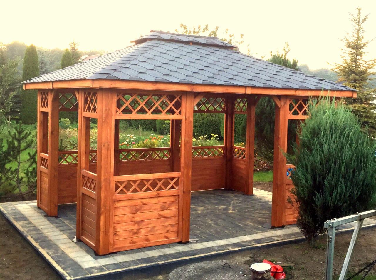 Drewniana altana z ciemnoszarym, łupkowym dachem, otoczona zielenią ogrodu, ustawiona na wybrukowanym tarasie.