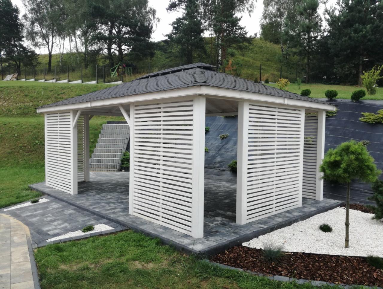 Biała altana z poziomymi listwami, ciemny dach, szara kostka brukowa i biały żwir w ogrodzie z zielonymi drzewami w tle.