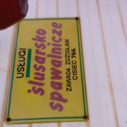 Żółta tabliczka informacyjna z napisem 'Usługi ślusarsko spawalnicze' oraz danymi kontaktowymi, umieszczona na ścianie z widocznym fragmentem rośliny w doniczce.