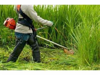 Osoba w roboczych ubraniach, z pomarańczowym kanistrem na plecach, przycina wysoką trawę za pomocą podkaszarki spalinowej.