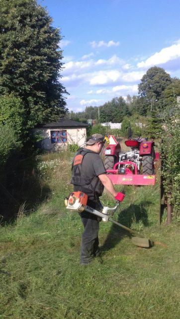 Mężczyzna w kamizelce i czapce kosi trawę za pomocą podkaszarki Stihl; w tle widoczny czerwony traktor z kosiarką rotacyjną, mały budynek i drzewa.