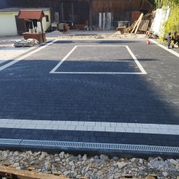 Dom-Bruk usługi budowlane - Nowo ułożona, wzorzysta nawierzchnia z ciemnej kostki brukowej z białymi pasami w Tochołowie, z widocznym odpływem liniowym i elementami krajobrazu w tle, częściowo w trakcie prac wykończeniowych.