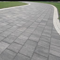 Dom-Bruk usługi budowlane - Szara, prostokątna nawierzchnia z kostki brukowej, obrzeża z drobnych kamieni, trawnik w tle, widok z perspektywy