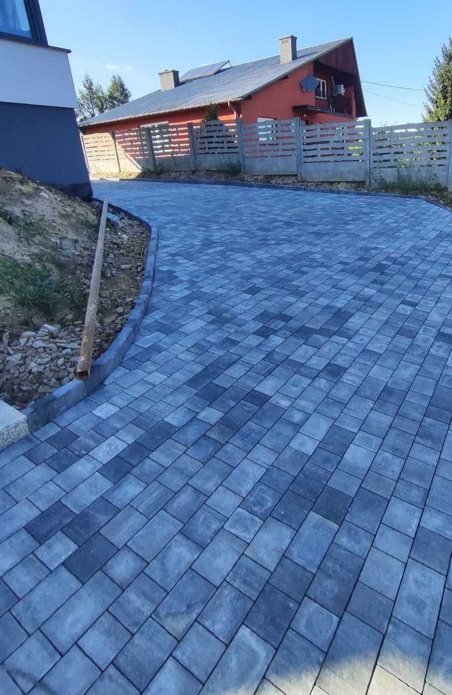 Ułożona z ciemnych i jasnych kostek brukowych ścieżka prowadząca do domu z szarym ogrodzeniem panelowym, widok z boku.