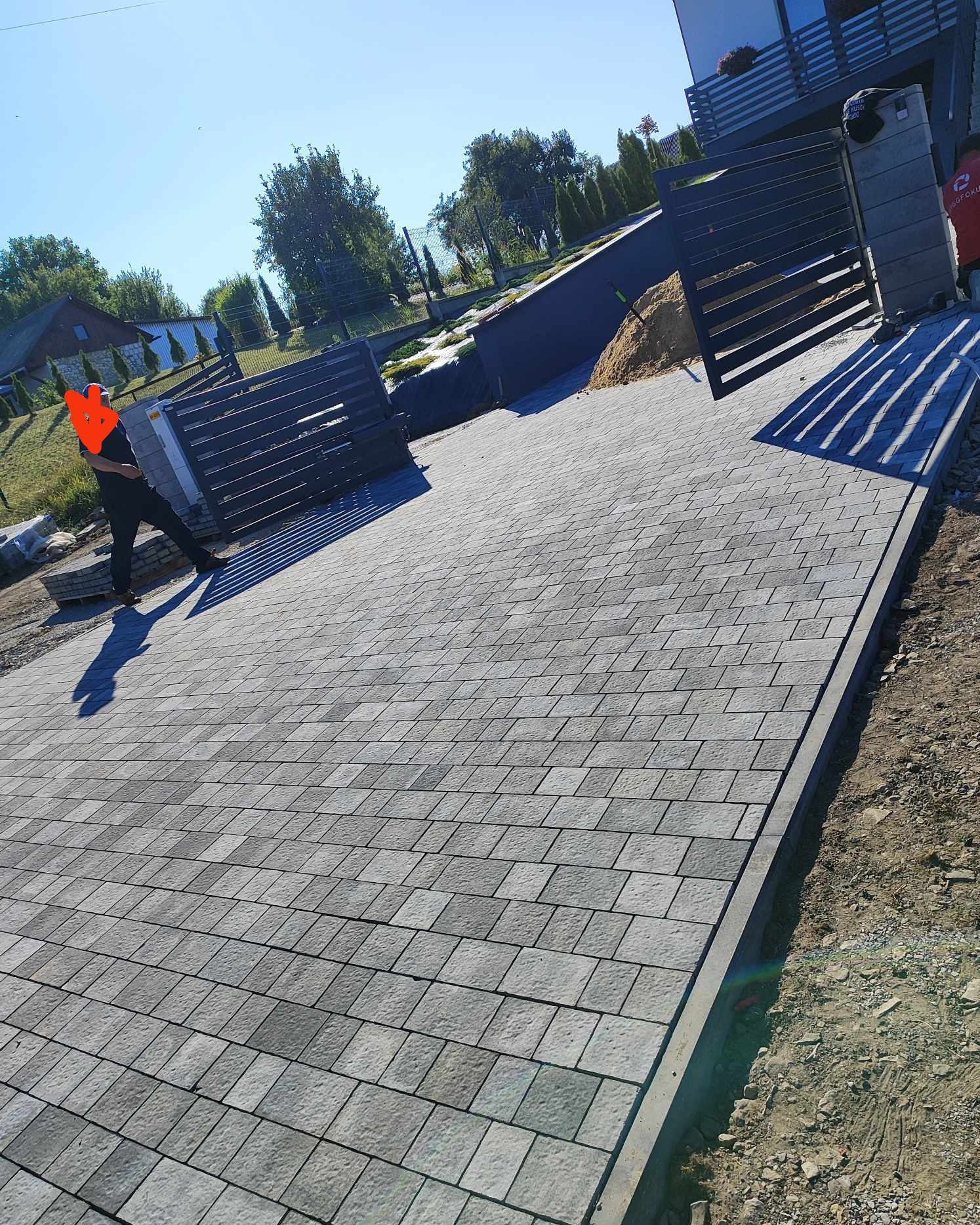 Ułożona szara kostka brukowa na podjeździe, widoczny pracownik w czarnym ubraniu, elementy ogrodzenia panelowego w tle, świecące słońce.