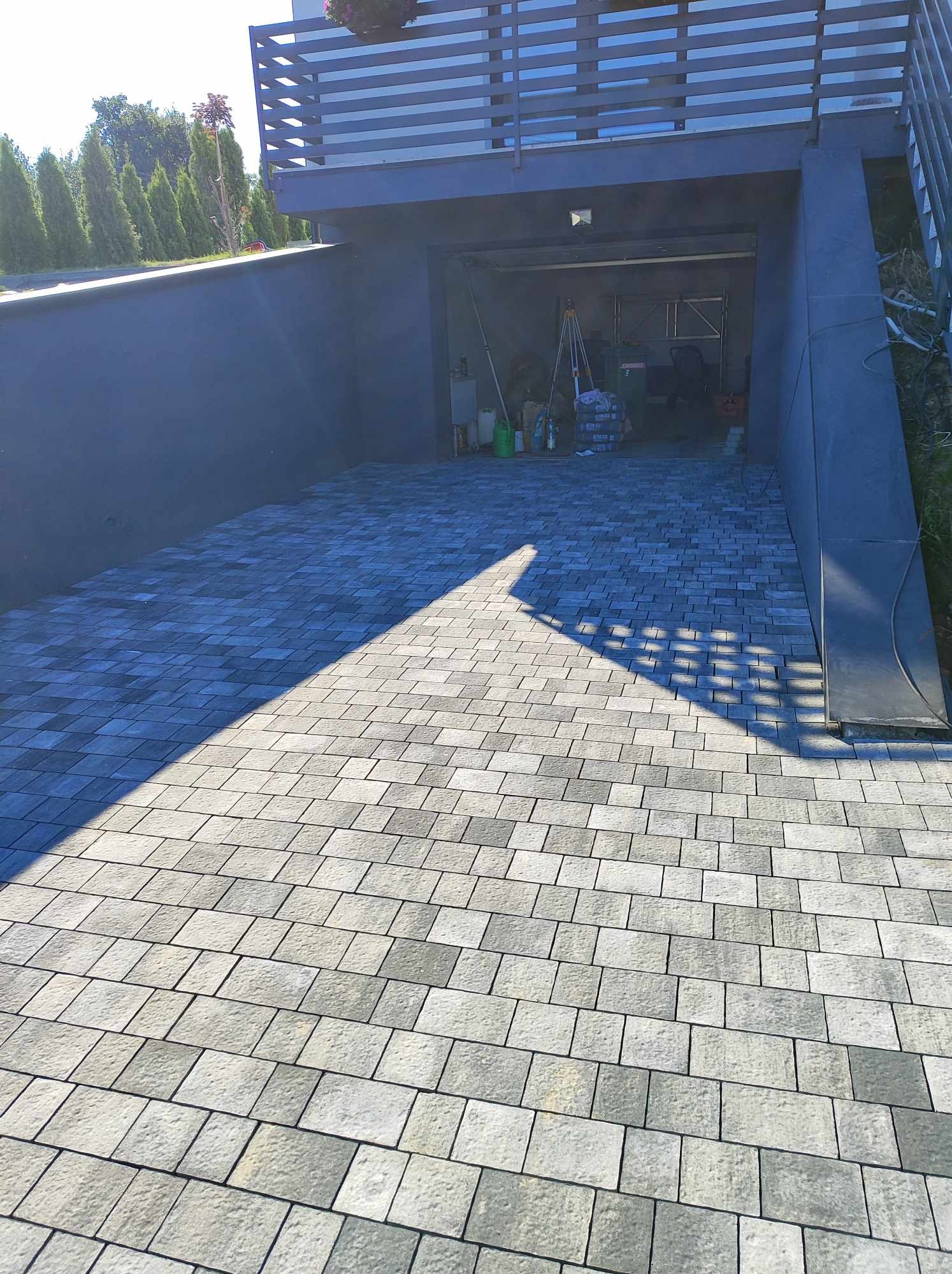 Ułożona szara kostka brukowa przed garażem w nowoczesnym domu z balkonem i zielonymi tujami w tle, cień rzucany na kostkę.
