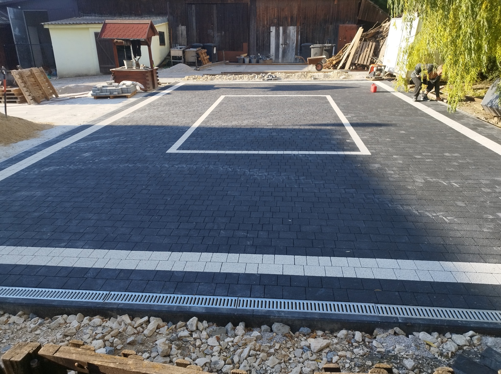 Nowo ułożona, wzorzysta nawierzchnia z ciemnej kostki brukowej z białymi pasami w Tochołowie, z widocznym odpływem liniowym i elementami krajobrazu w tle, częściowo w trakcie prac wykończeniowych.