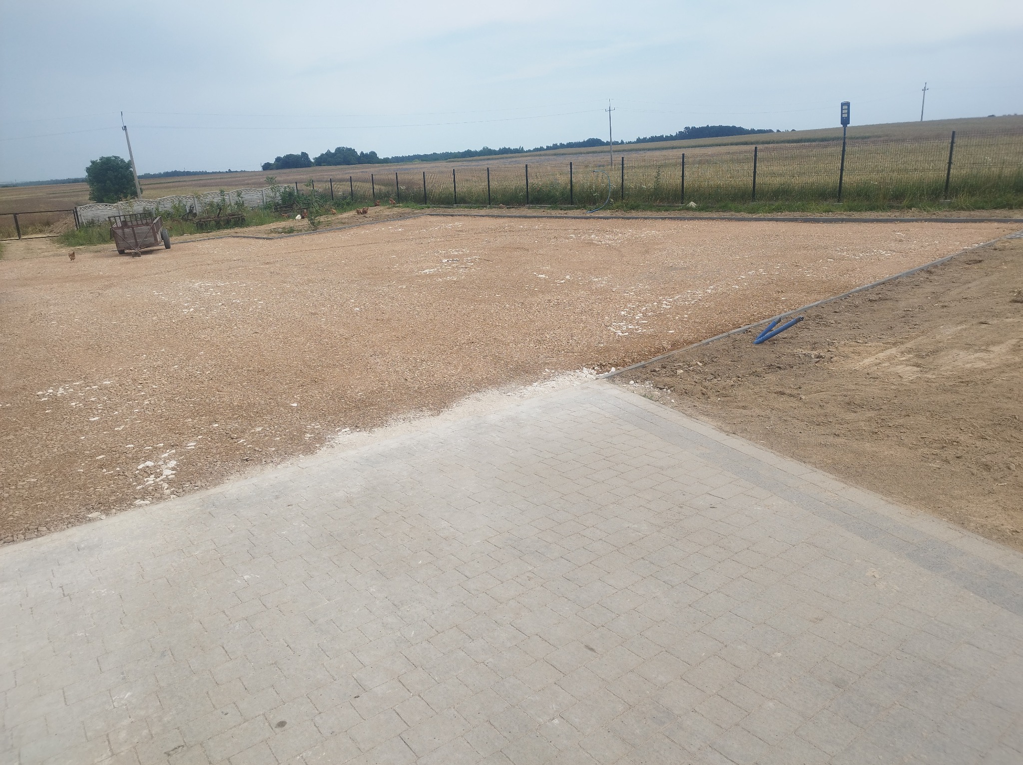 Ukończona nawierzchnia z kostki brukowej przechodząca w teren utwardzony kruszywem, widoczne ogrodzenie panelowe w tle i wiejski krajobraz.