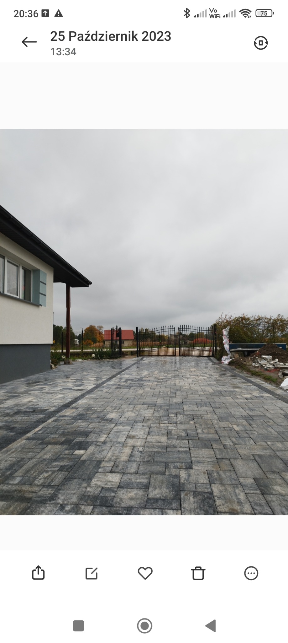 Szara kostka brukowa ułożona na podjeździe przed domem z czarną bramą w tle, widok na pochmurne niebo.