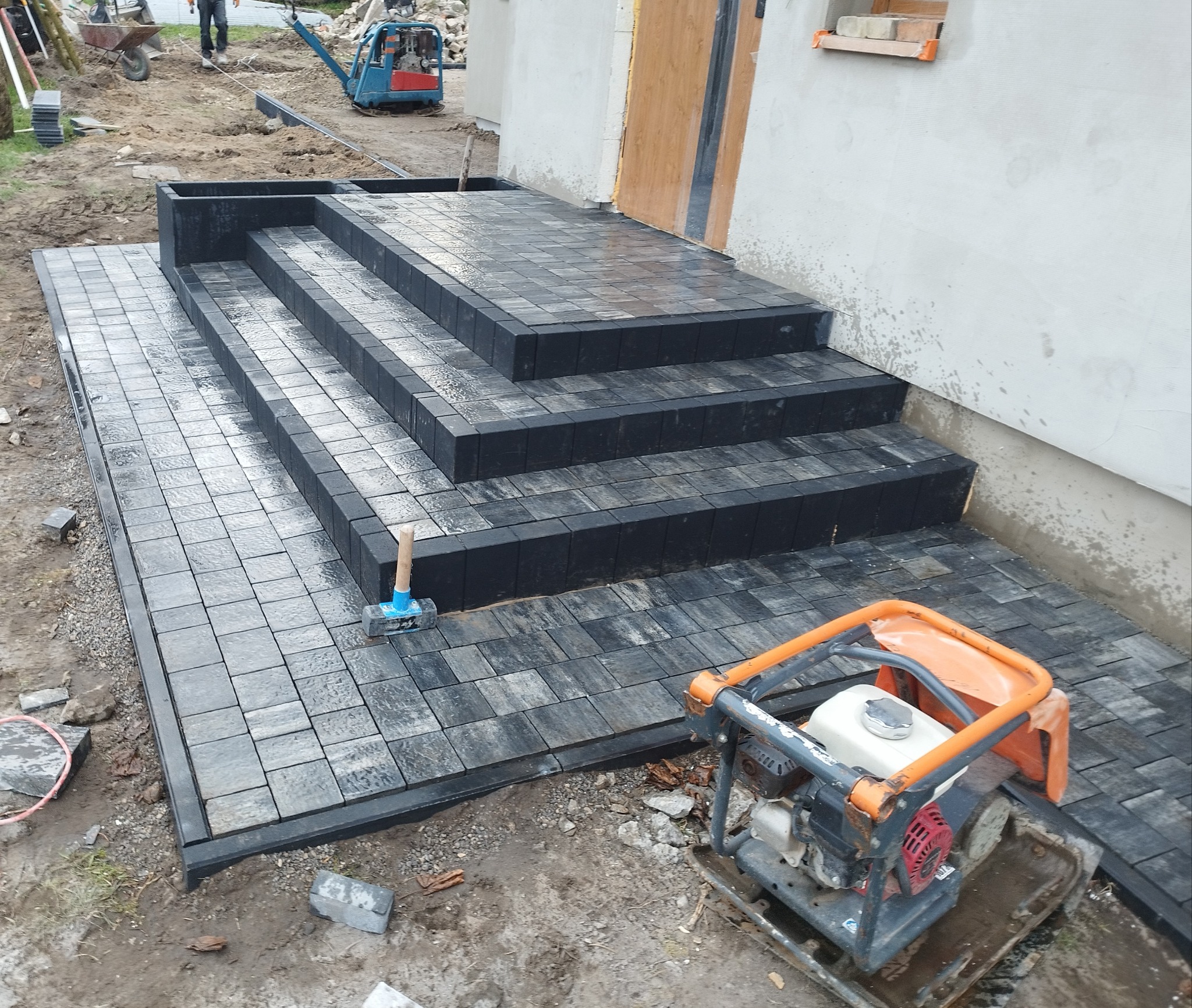 Ułożone schody z kostki brukowej w kolorze szarym, prowadzące do drzwi budynku, z widocznym ubijakiem wibracyjnym i gumowym młotkiem brukarskim.