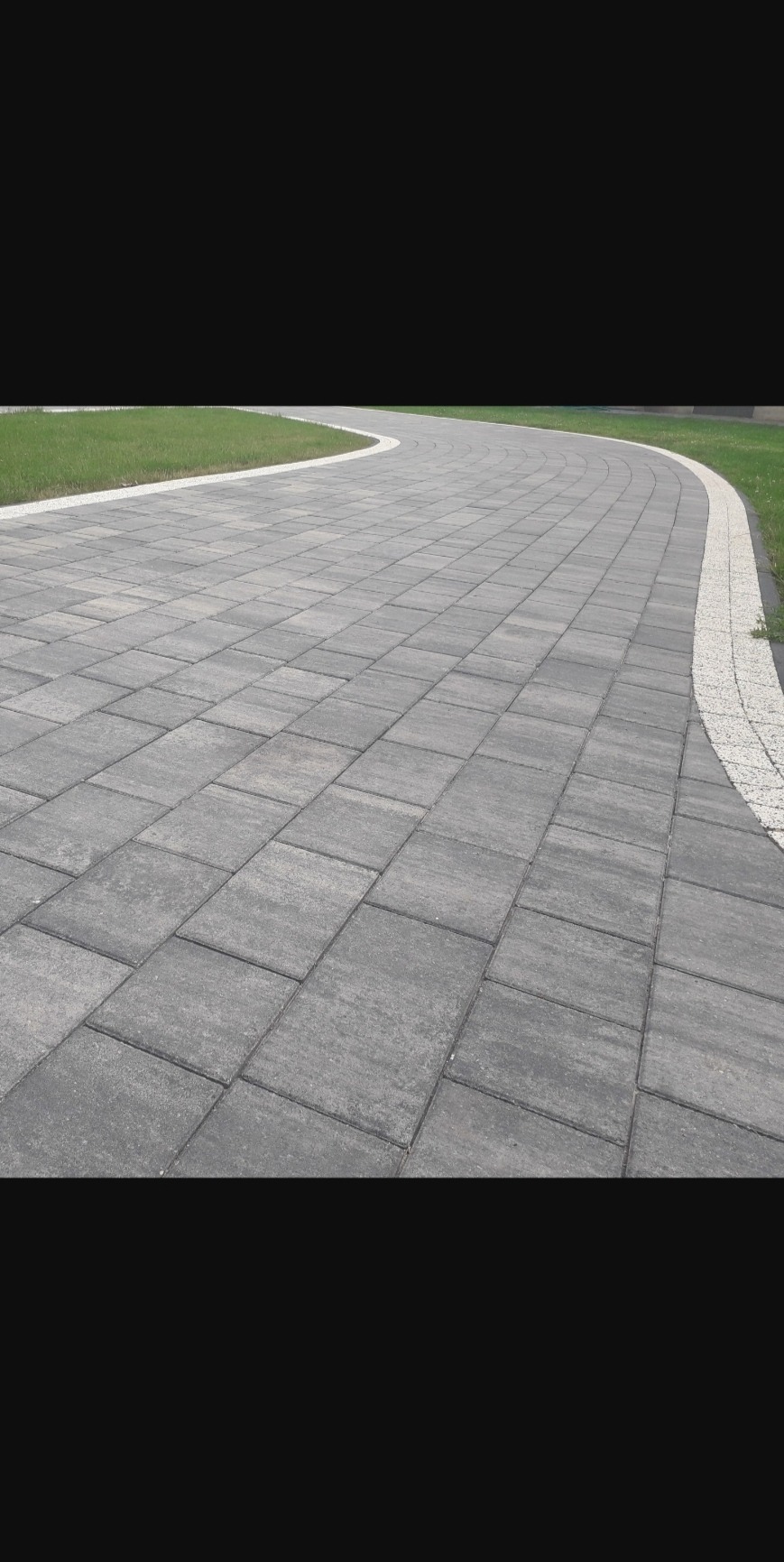 Szara, prostokątna nawierzchnia z kostki brukowej, obrzeża z drobnych kamieni, trawnik w tle, widok z perspektywy