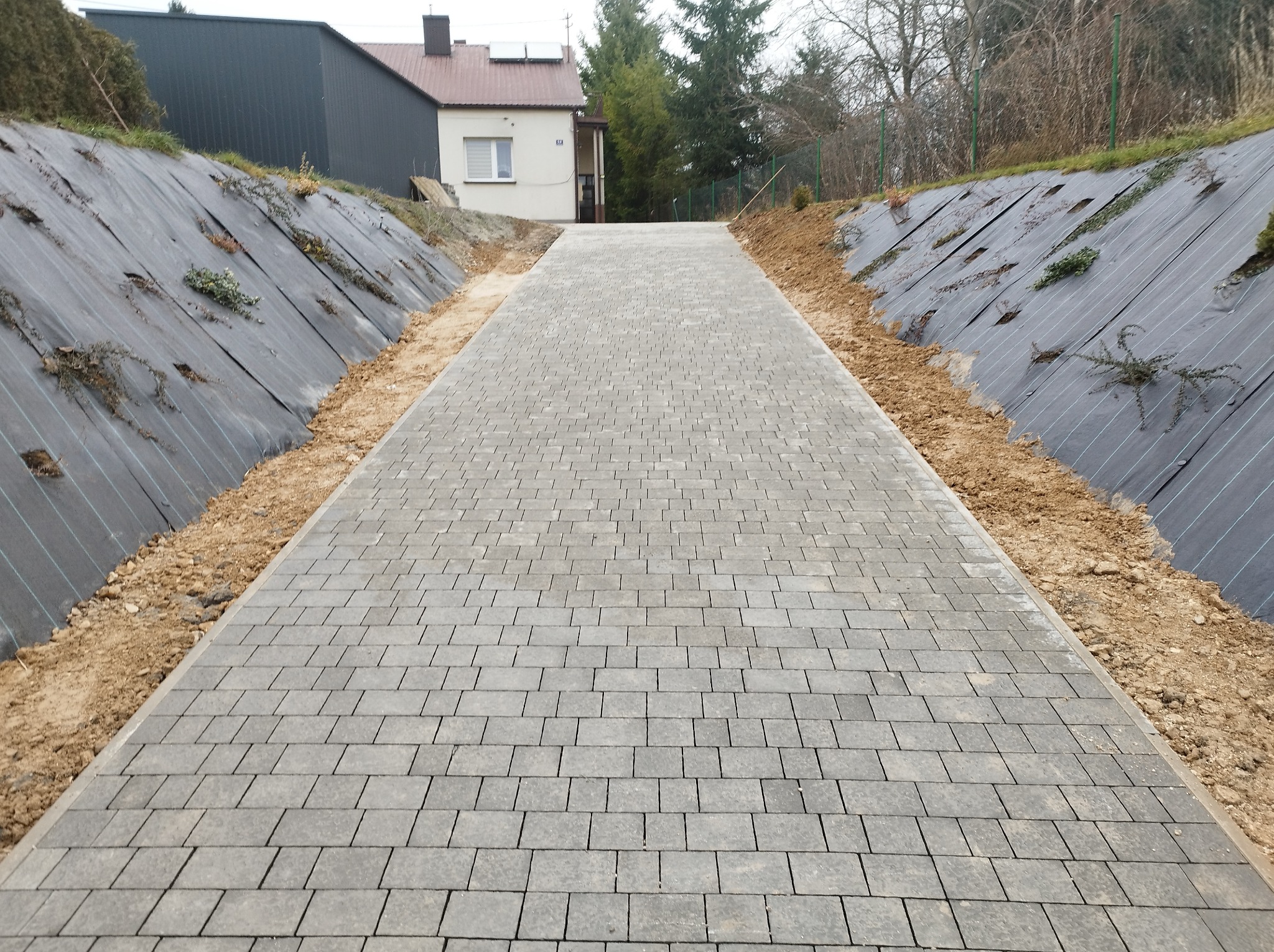 Szara ścieżka z kostki prowadząca pod górę w kierunku domu, po bokach skarpy zabezpieczone czarną agrowłókniną, częściowo porośniętą roślinnością.