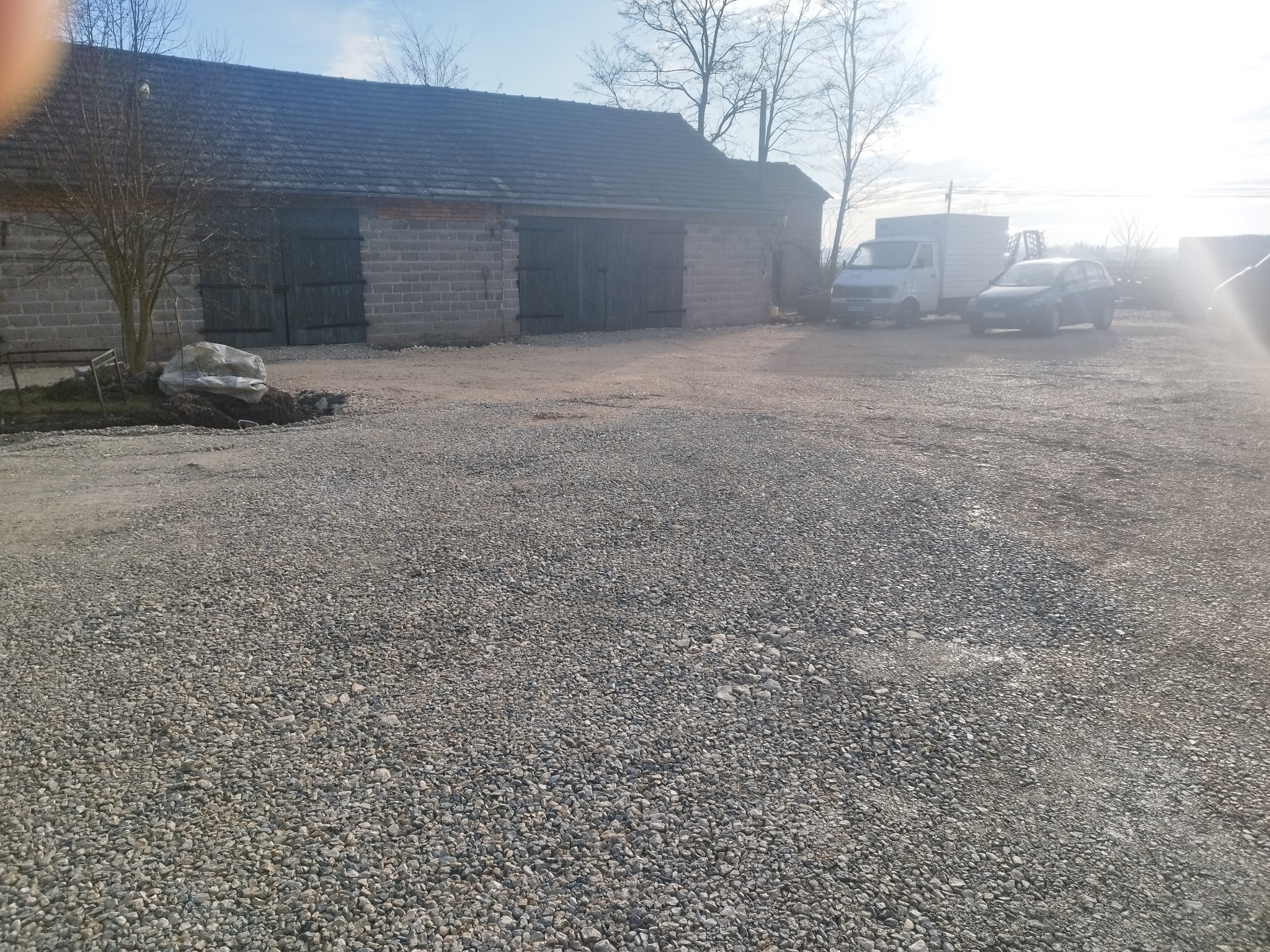 Szeroki plan żwirowej powierzchni przed budynkiem gospodarczym z dwoma bramami, w tle zaparkowane samochody dostawcze i osobowe, drzewa bez liści, jasne, słoneczne oświetlenie.