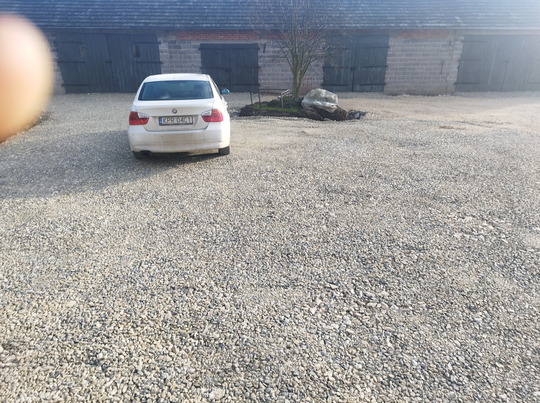 Biały samochód osobowy zaparkowany na szutrowym podwórku przed budynkiem gospodarczym z szarych cegieł z drewnianymi wrotami, w tle małe drzewko i kamień.