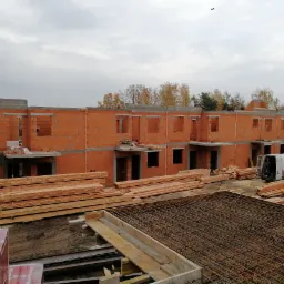 Budowa szeregu domów jednorodzinnych w stanie surowym otwartym, widoczne ściany z czerwonej cegły, przygotowane zbrojenie fundamentów i stos drewna budowlanego, biały bus dostawczy na placu budowy,...