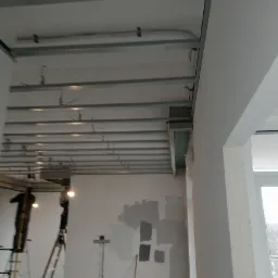 Montaż metalowego rusztu podwieszanego sufitu z płyty gipsowo-kartonowej, widoczne profile CD i UD, instalacje elektryczne, ściany przygotowane do malowania, drabiny w tle.