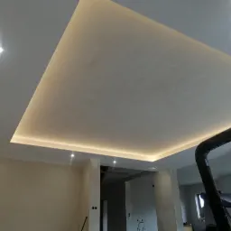 Nowoczesny sufit podwieszany z oświetleniem LED w kształcie trapezu, widoczne punktowe lampy halogenowe i fragment instalacji wentylacyjnej, wnętrze w trakcie prac wykończeniowych.