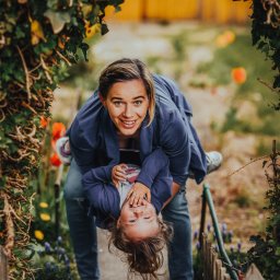 Fotoxfoto.pl Krzysztof Sobczyński - Uśmiechnięta mama trzymająca córkę do góry nogami na tle ogrodu z kwitnącymi tulipanami i łukiem z bluszczu.