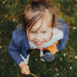 Fotoxfoto.pl Krzysztof Sobczyński - Dziewczynka z brązowymi włosami spiętymi w kok, uśmiecha się patrząc w górę, trzymając w ręku lizaka na tle zielonej trawy, ujęcie z góry.