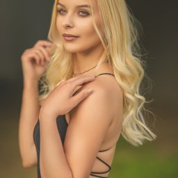 Fotoxfoto.pl Krzysztof Sobczyński - Portret młodej kobiety z blond włosami, ubrana w czarną sukienkę z odkrytymi plecami, delikatny makijaż, spogląda w bok, dłoń na ramieniu, złoty naszyjnik.