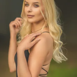 Portret młodej kobiety z blond włosami, ubrana w czarną sukienkę z odkrytymi plecami, delikatny makijaż, spogląda w bok, dłoń na ramieniu, złoty naszyjnik.