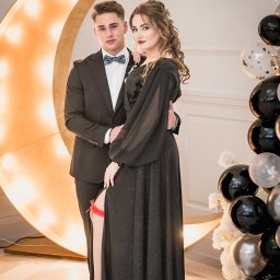 Fotoxfoto.pl Krzysztof Sobczyński - Elegancka para pozuje na studniówce: on w czarnym garniturze i muszce, ona w długiej, błyszczącej sukni z rozcięciem, eksponując podwiązkę, na tle dekoracji z podświetlanego księżyca i balonów.