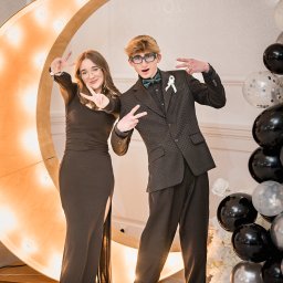 Fotoxfoto.pl Krzysztof Sobczyński - Dwoje nastolatków w eleganckich strojach pozuje z gestem zwycięstwa na tle dekoracji z żarówkami w kształcie księżyca i balonów w Jarosławiu, wykonane przez Fotoxfoto.pl.