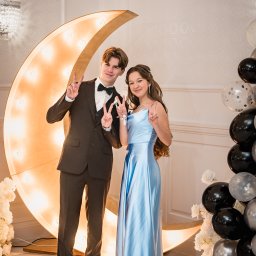 Fotoxfoto.pl Krzysztof Sobczyński - Uśmiechnięta para w strojach wieczorowych pozuje na tle dekoracji z podświetlanego księżyca i balonów podczas studniówki.