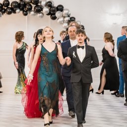 Fotoxfoto.pl Krzysztof Sobczyński - Para prowadzi poloneza na studniówce, dziewczyna w zielonej sukience z koronkowymi aplikacjami, chłopak w czarnym garniturze z muszką, w tle łuk z czarno-srebrnych balonów i rzędy gości w strojach...