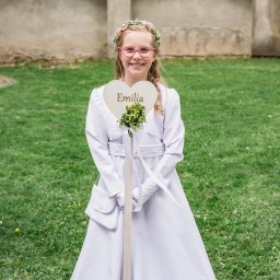 Fotoxfoto.pl Krzysztof Sobczyński - Uśmiechnięta dziewczynka w białej sukience komunijnej, z wiankiem i tabliczką z imieniem 'Emilia', na tle zielonego trawnika i muru z ceglanym dachem.