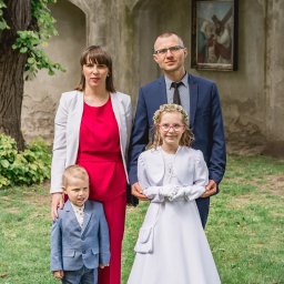 Fotoxfoto.pl Krzysztof Sobczyński - Uroczysta fotografia rodzinna w plenerze: rodzice z dziećmi, dziewczynka w albie komunijnej, chłopiec w garniturze. W tle budynek z obrazem religijnym w niszy.