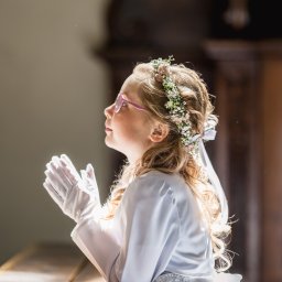 Fotoxfoto.pl Krzysztof Sobczyński - Dziewczynka w białej sukience komunijnej z wiankiem na głowie i różowych okularach, modli się w kościele, oświetlona promieniami słońca. Profil, dłonie złożone.