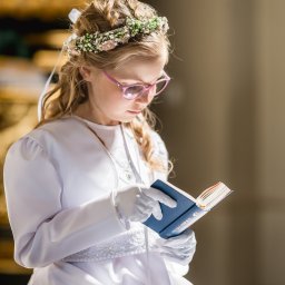Fotoxfoto.pl Krzysztof Sobczyński - Dziewczynka w białej albie i wianku czyta modlitewnik w kościele, podczas uroczystości pierwszej komunii. Delikatne światło, w tle zarys wnętrza.