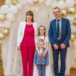 Uśmiechnięta rodzina pozuje na tle dekoracji z białych i złotych balonów podczas uroczystości Pierwszej Komunii Świętej. Mama w różowym, tata w niebieskim garniturze, dzieci w bieli i błękicie.